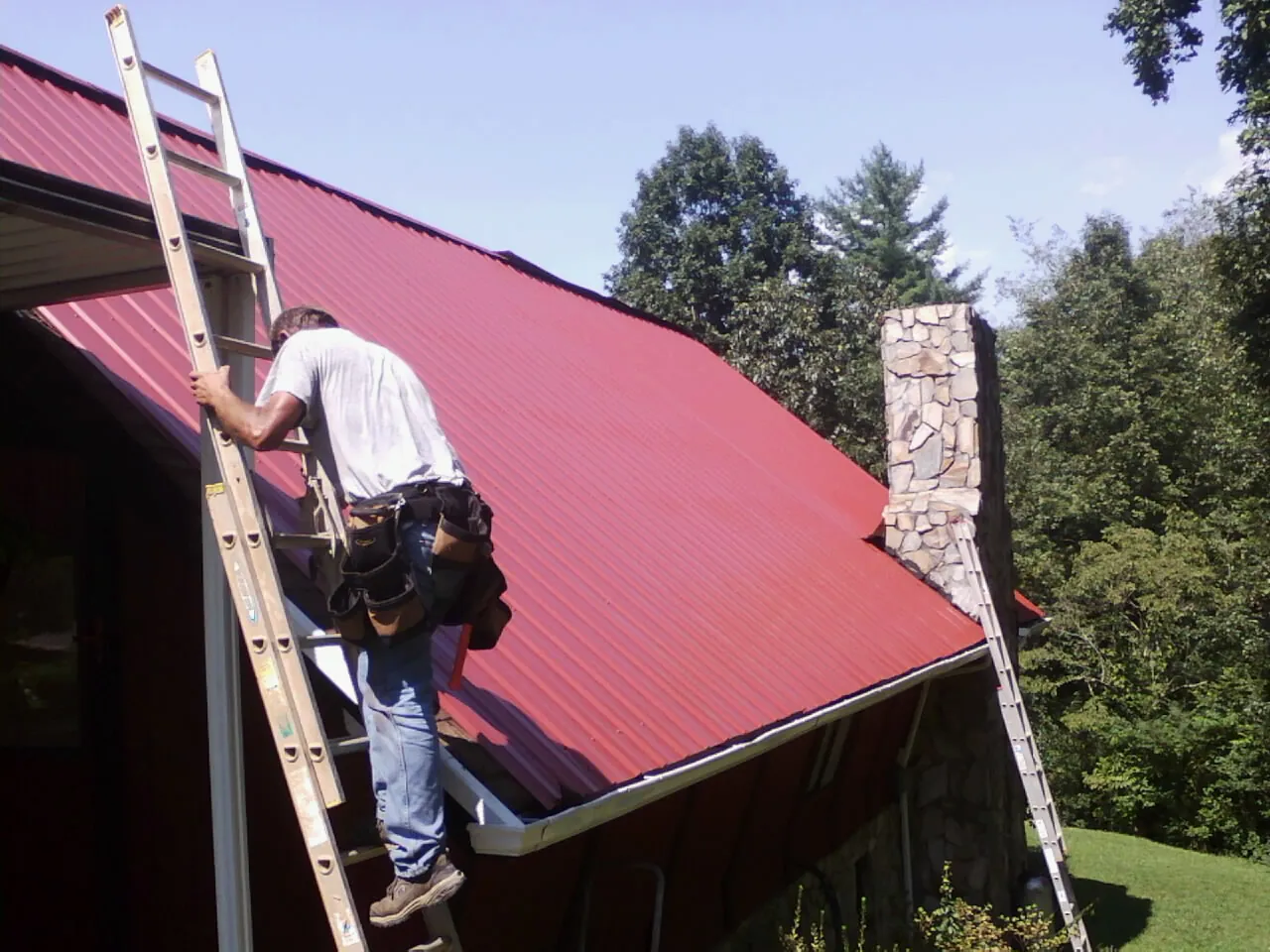 Roofer on a ladder beside a red metal roof and stone chimney — Soffit & Fascia Repair by Roofing Brothers in Silver Gate